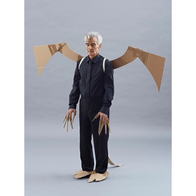 Full shot of a man in a black shirt and pants wearing a cardboard dragon costume with wings, horns, claws, and feet against a grey backdrop.