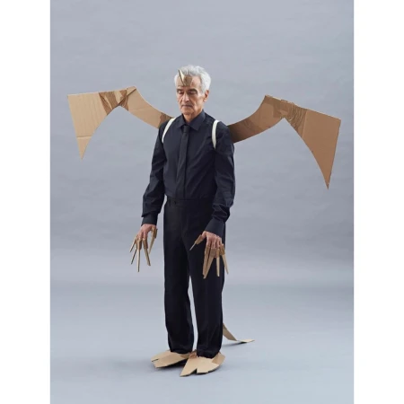 Full shot of a man in a black shirt and pants wearing a cardboard dragon costume with wings, horns, claws, and feet against a grey backdrop.