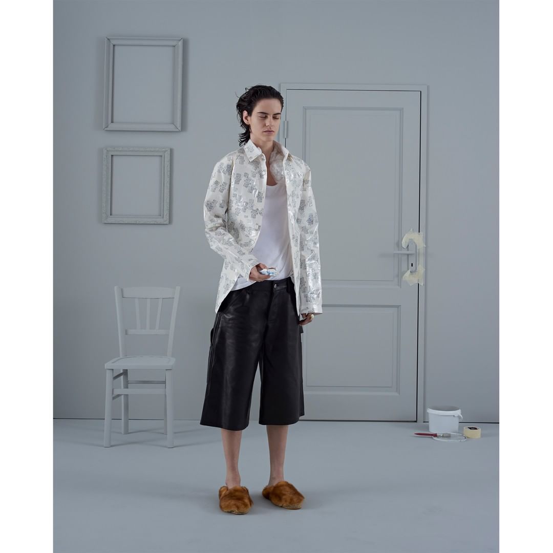 Studio shot of a person in a white floral jacket, black shorts, and furry slippers holding a small device, standing in a gray room.
