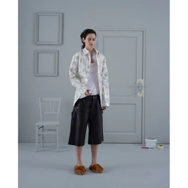 Studio shot of a person in a white floral jacket, black shorts, and furry slippers holding a small device, standing in a gray room.
