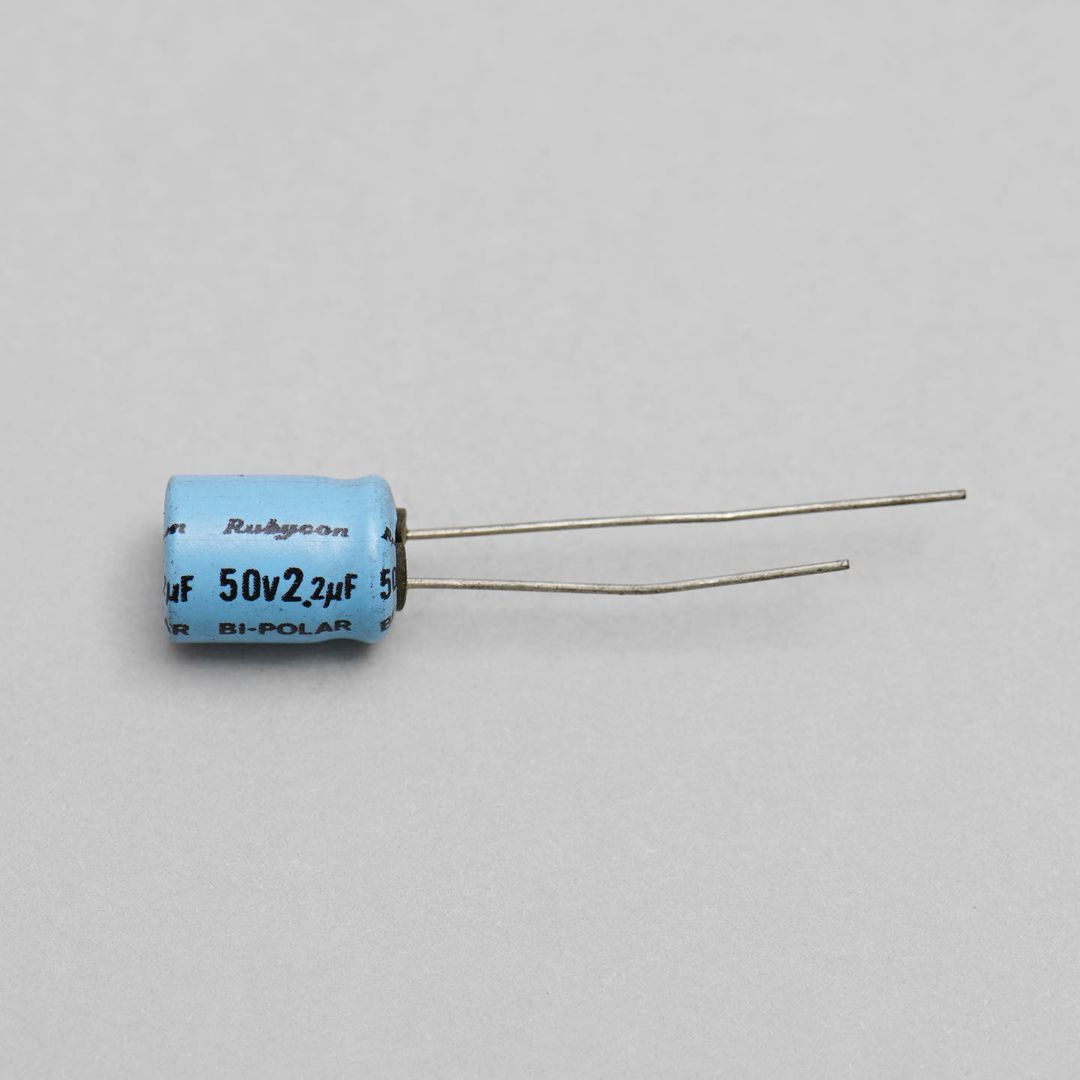 Close-up product shot of a light blue Rubycon capacitor with silver wires against a grey background. Electronic component detail.