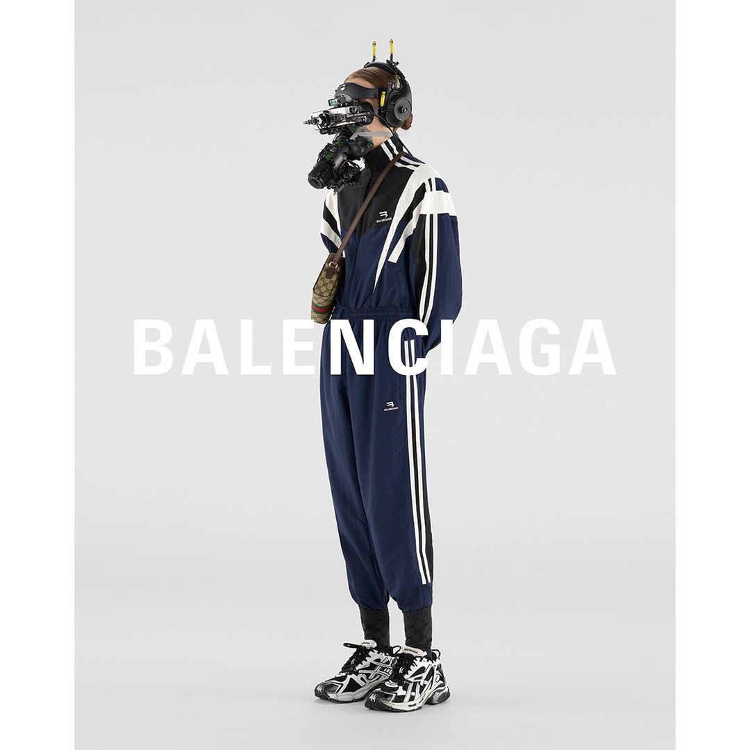 Full-length studio shot of a person wearing a navy and white track suit and futuristic-looking headgear against a white backdrop.