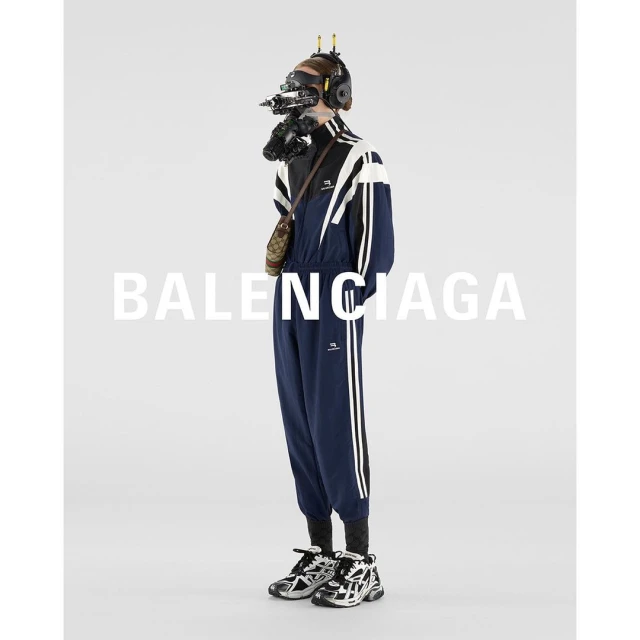 Full-length studio shot of a person wearing a navy and white track suit and futuristic-looking headgear against a white backdrop.