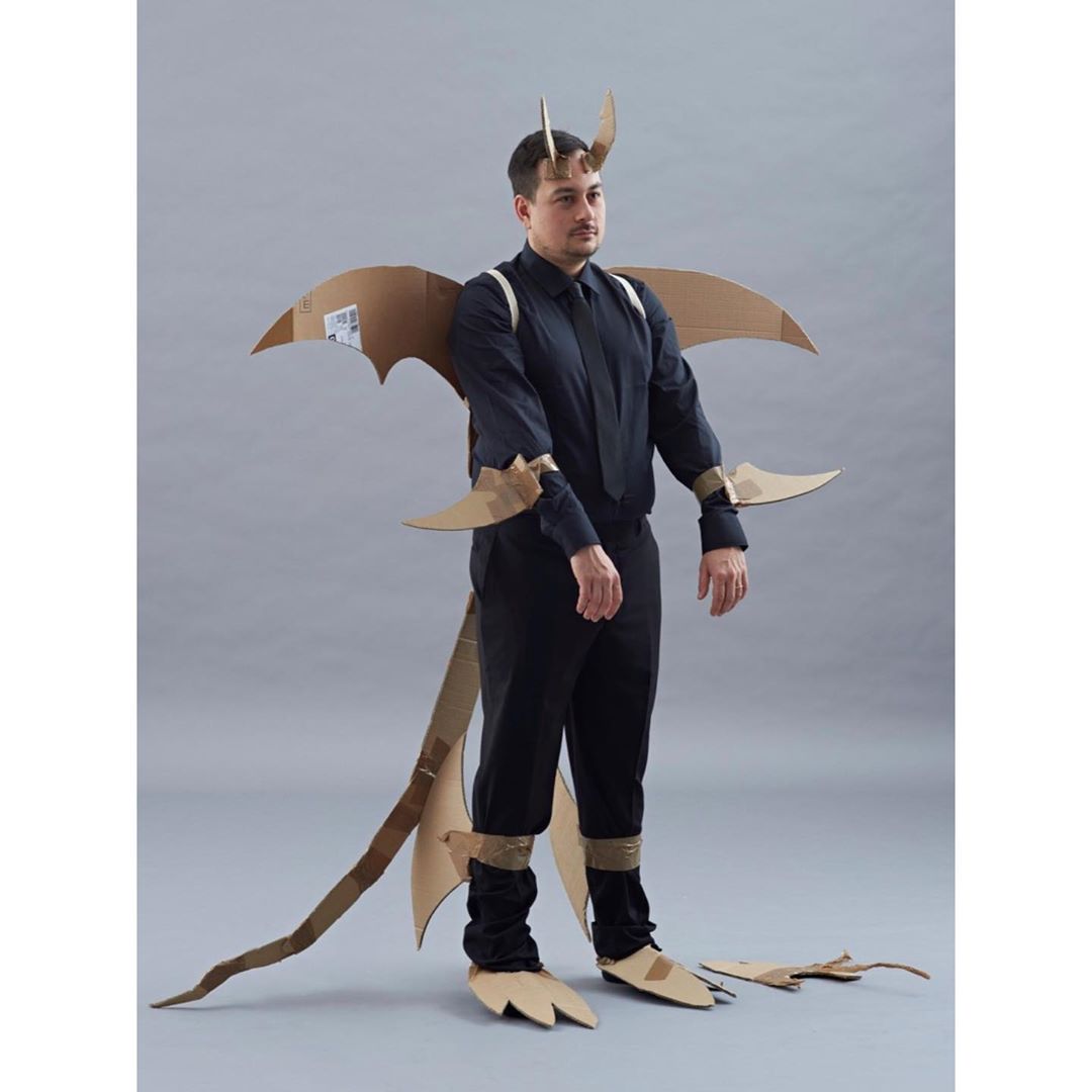 Man in a cardboard dragon costume with wings, tail, horns, and clawed feet, posing against a grey background, wearing black shirt and pants.
