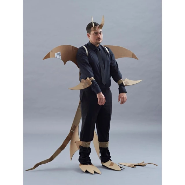 Man in a cardboard dragon costume with wings, tail, horns, and clawed feet, posing against a grey background, wearing black shirt and pants.