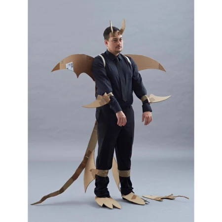 Man in a cardboard dragon costume with wings, tail, horns, and clawed feet, posing against a grey background, wearing black shirt and pants.