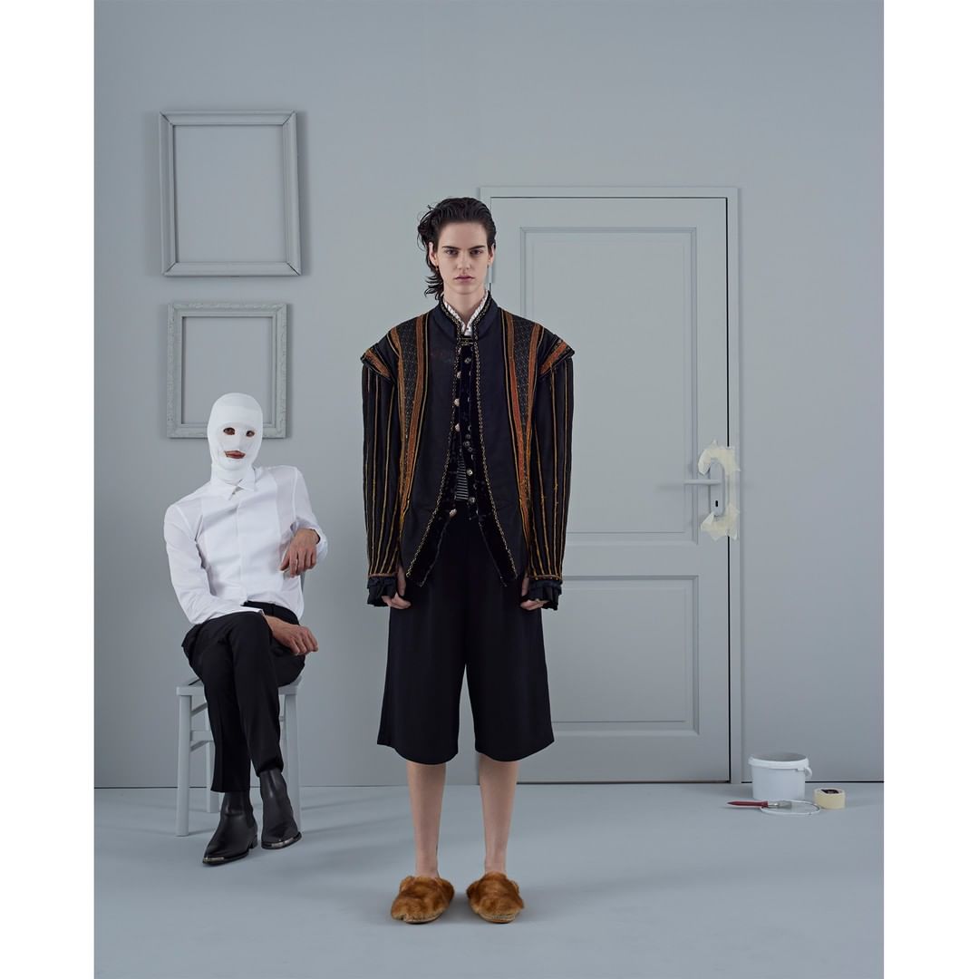 Two figures in a grey room; one standing in a black suit with an ornate jacket and one seated wearing a white mask and button down shirt.