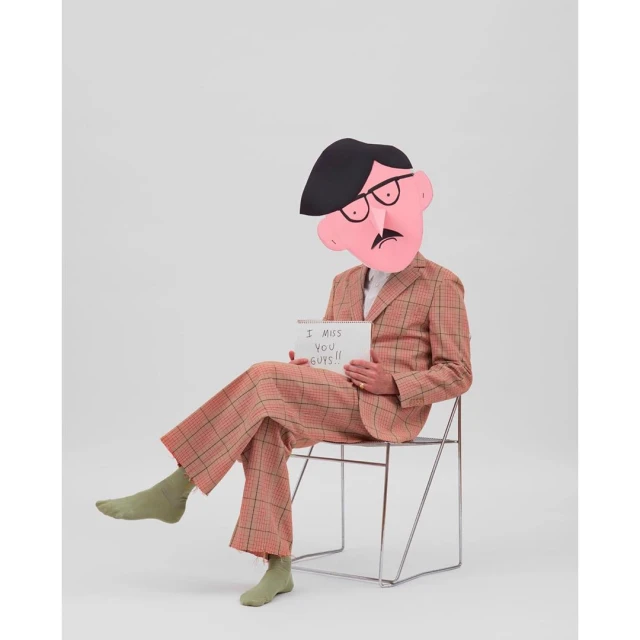 Person in a plaid suit sits on a wire chair, face obscured by a cartoon head, holding a sign that reads, 'I miss you guys!!'