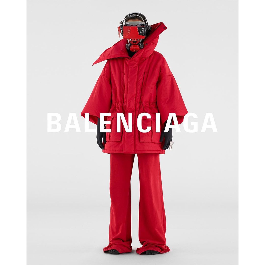 A person wearing red oversized outerwear and headgear stands in a white studio, featuring the Balenciaga logo in the center.