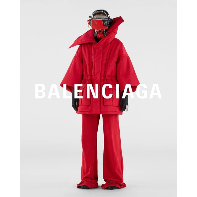 A person wearing red oversized outerwear and headgear stands in a white studio, featuring the Balenciaga logo in the center.