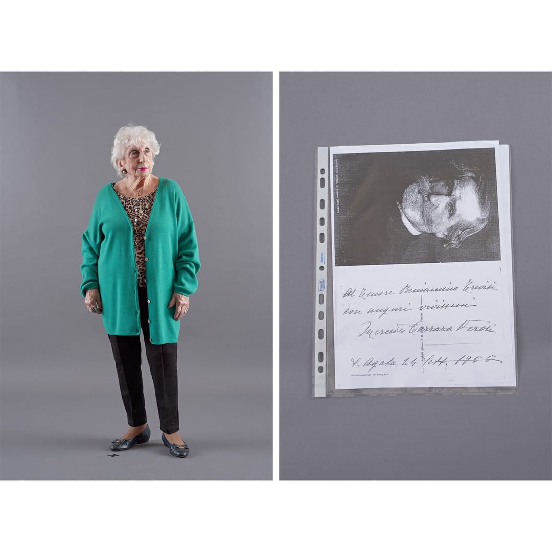 Full-length shot of an elderly woman in a green cardigan and black pants standing next to a document on gray background.