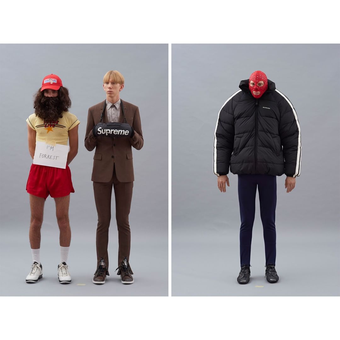 Three figures standing in front of a grey wall, two costumed: one like Forrest Gump holding a sign, and another in a mask.