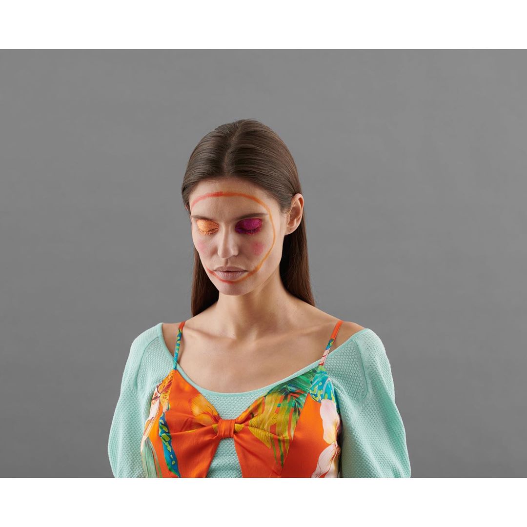 Woman with creative makeup, wearing a colorful floral top and light-green sweater, poses in a studio with a neutral gray backdrop.