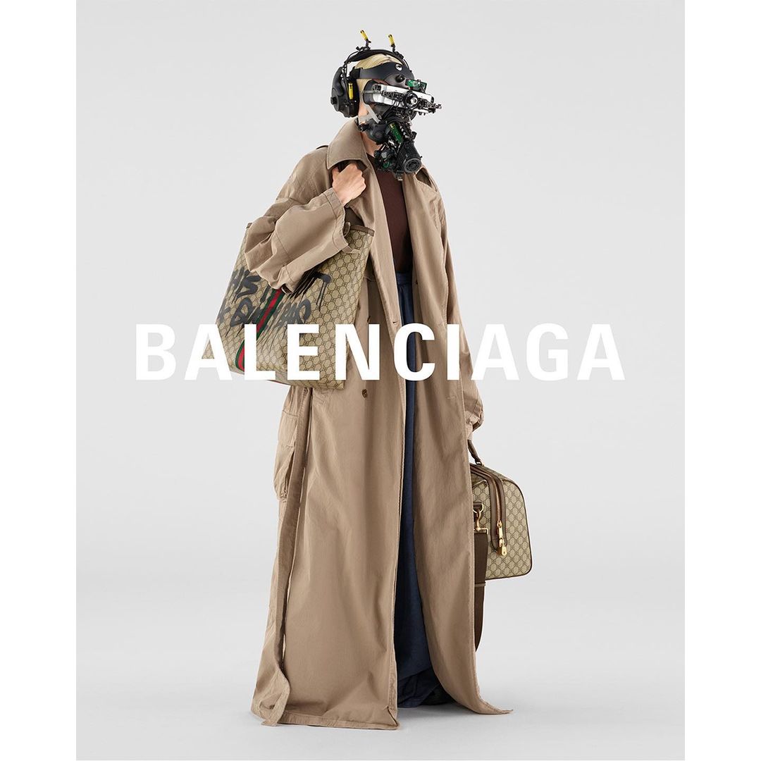 Model with sci-fi helmet, tan trench coat, and logo bags in a neutral studio setting. Fashion-forward and technologically infused editorial shot.
