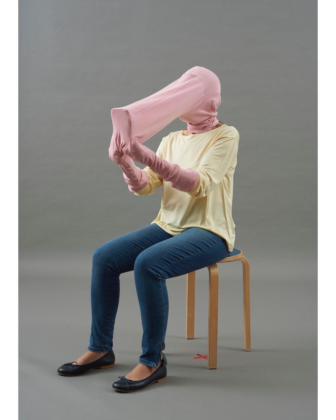 Person sitting on stool wearing pink head covering, matching long gloves, blue jeans, yellow top, black shoes against gray background.