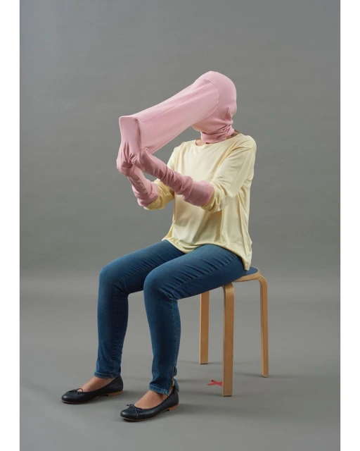 Person sitting on stool wearing pink head covering, matching long gloves, blue jeans, yellow top, black shoes against gray background.
