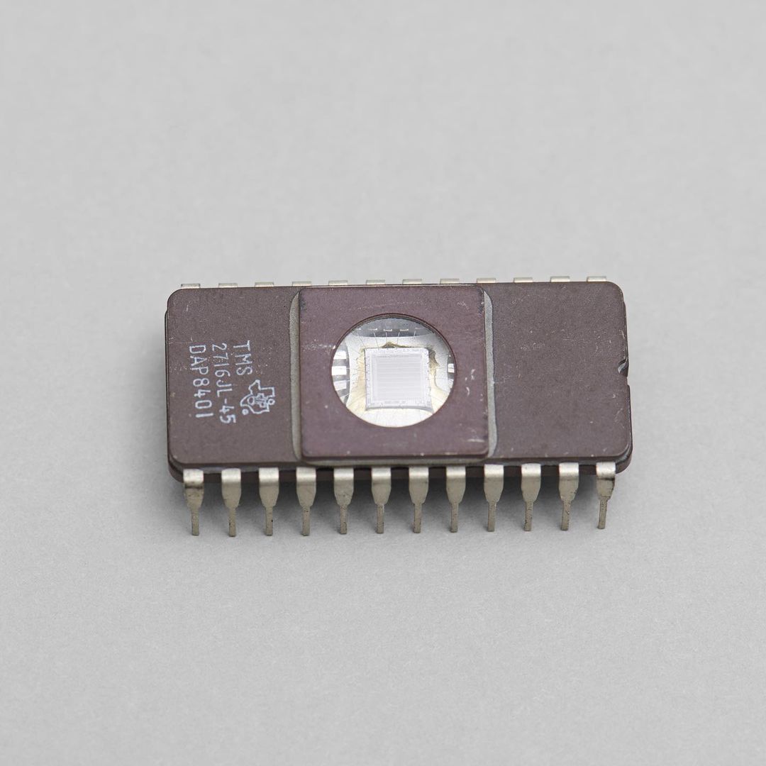 Brown microchip with metal pins and a transparent square exposing the chip's circuitry, photographed in a close-up shot against a grey background.