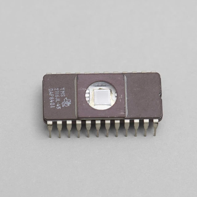Brown microchip with metal pins and a transparent square exposing the chip's circuitry, photographed in a close-up shot against a grey background.