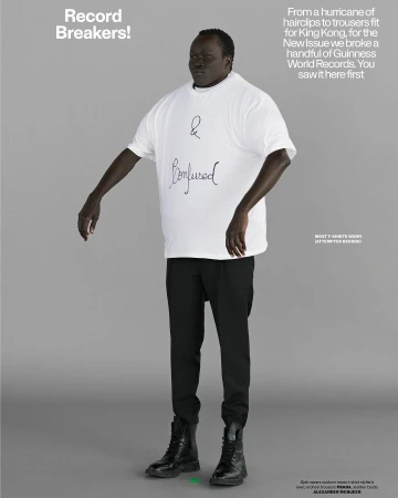 A tall, dark-skinned man stands centered in a full shot, wearing a white '& Confused' t-shirt, black pants, and boots against a neutral backdrop.