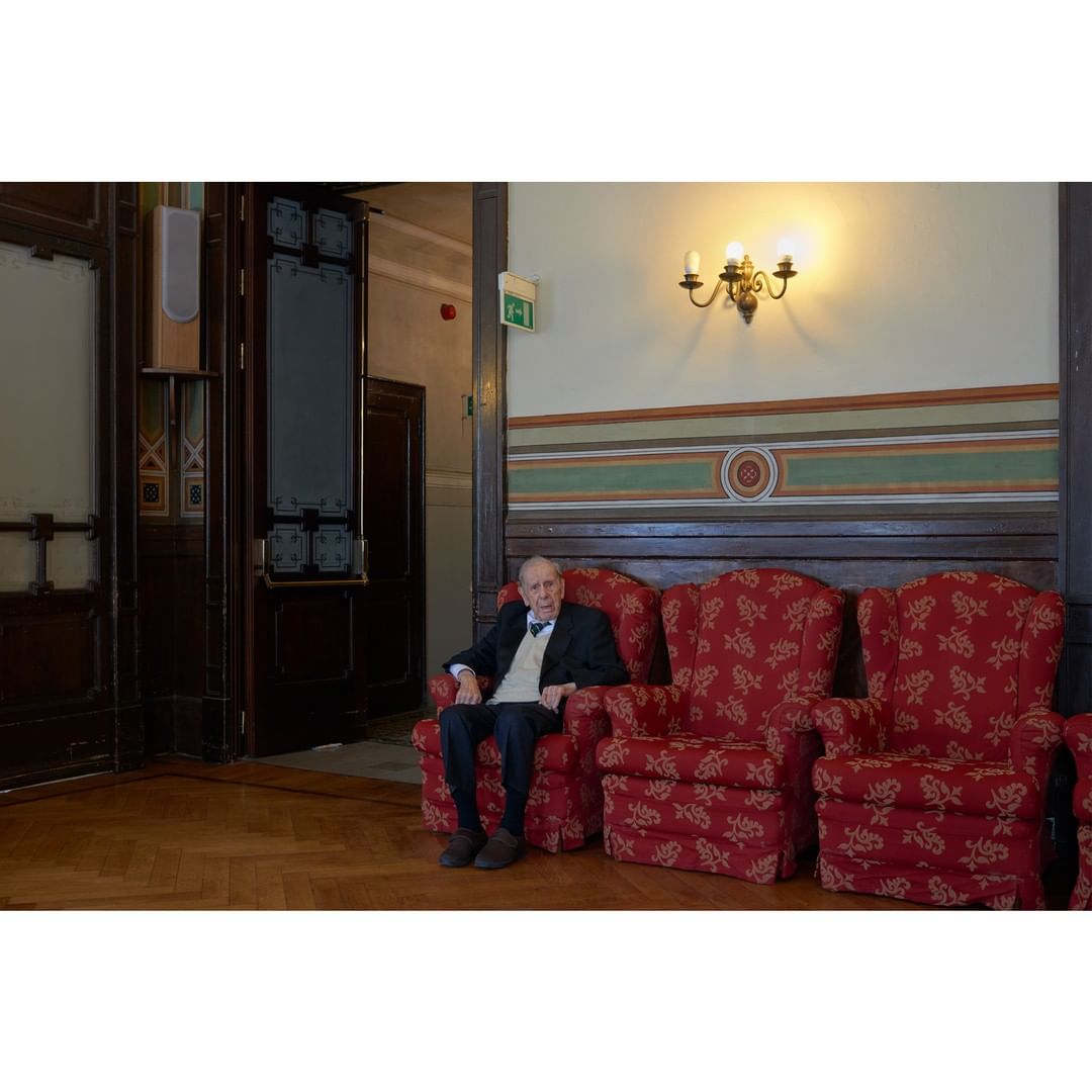 Elderly man seated in a patterned armchair indoors, wearing a dark suit and light vest, in a hall with ornate woodwork.
