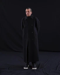Man wearing a long black double-breasted coat standing against a dark seamless background