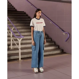 Young woman standing on a tiled floor wearing a white ringer t-shirt, blue cargo jeans, and platform shoes near stairs with a purple handrail.