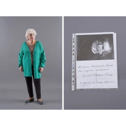Elderly woman standing in a green cardigan next to a plastic-sleeved document on a grey background