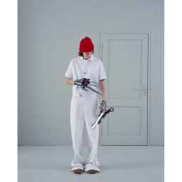 Model in white polo shirt, red beanie, and white trousers holding metallic claw props in a grey studio setting.