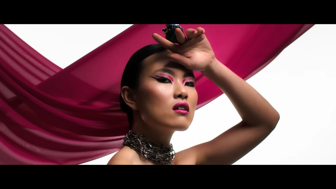Close-up of a stylish woman with pink eye makeup, glossy lipstick, and a statement chain. A sheer scarf frames her face.