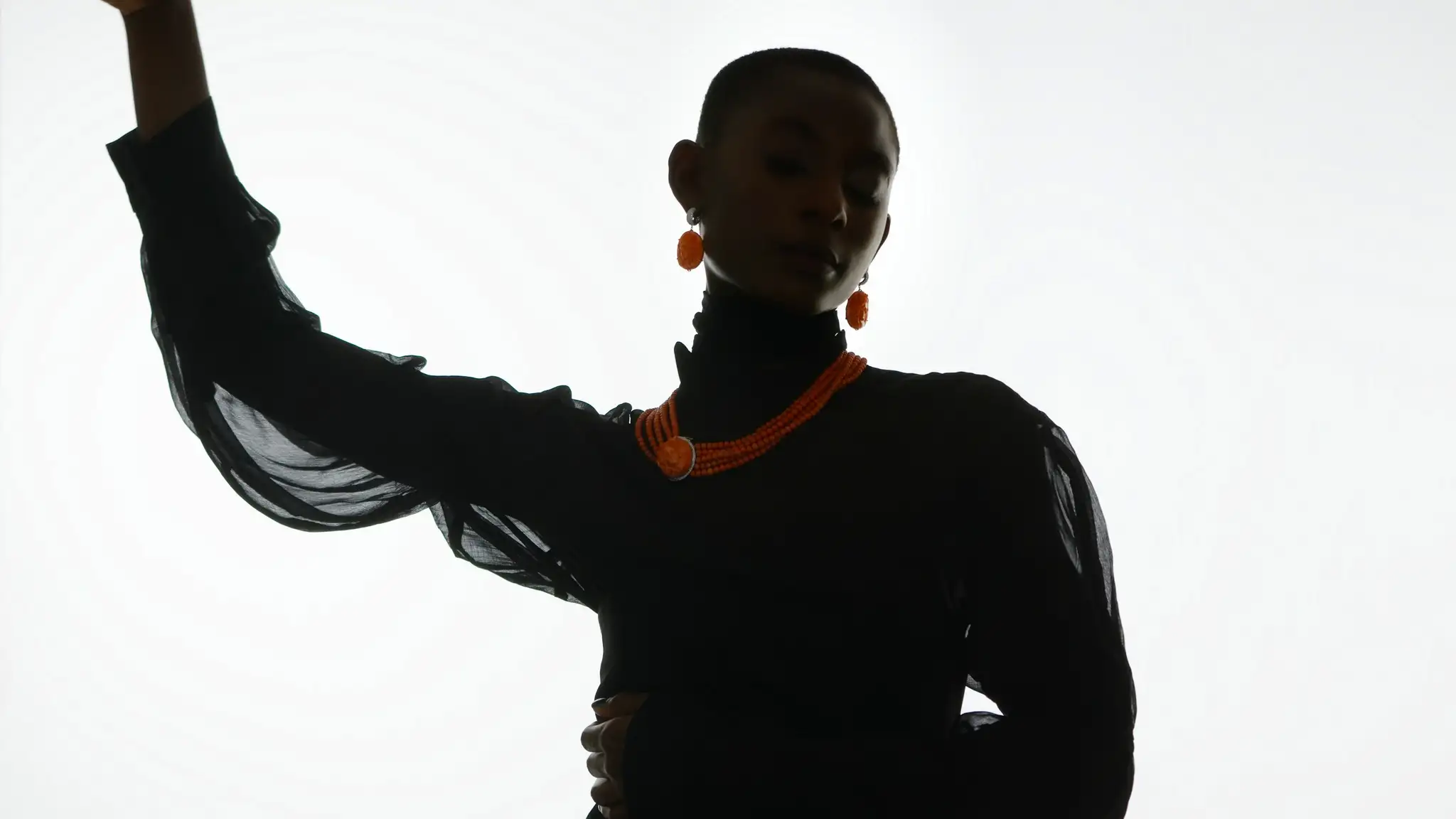 Fashionable woman with short hair poses in a sheer black long-sleeve turtleneck, orange necklace and matching earrings on white backdrop.