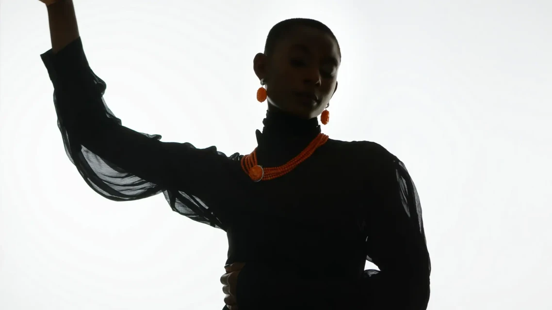 Fashionable woman with short hair poses in a sheer black long-sleeve turtleneck, orange necklace and matching earrings on white backdrop.
