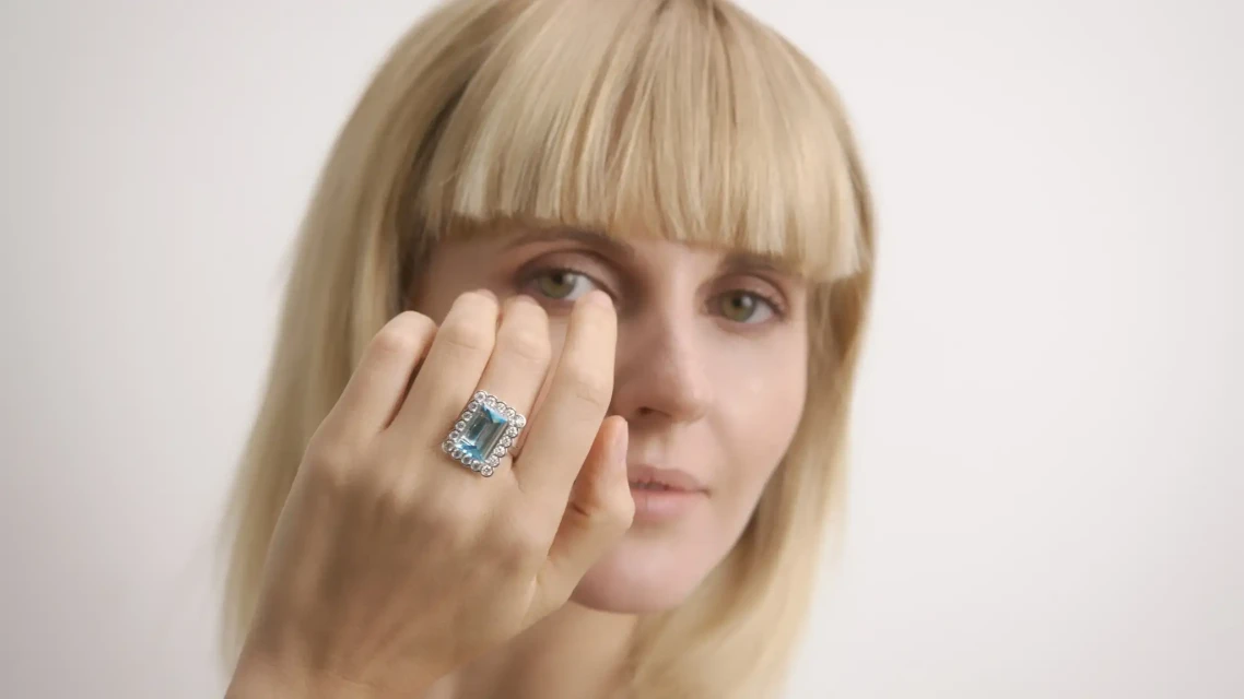 Woman with blonde bangs poses indoors wearing a large gemstone ring; focus on the jewelry and her neutral expression.