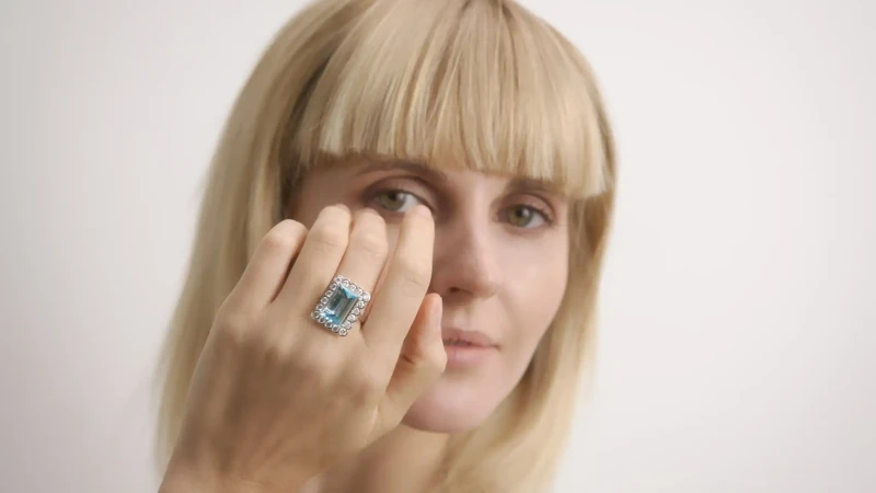 Woman with blonde bangs poses indoors wearing a large gemstone ring; focus on the jewelry and her neutral expression.