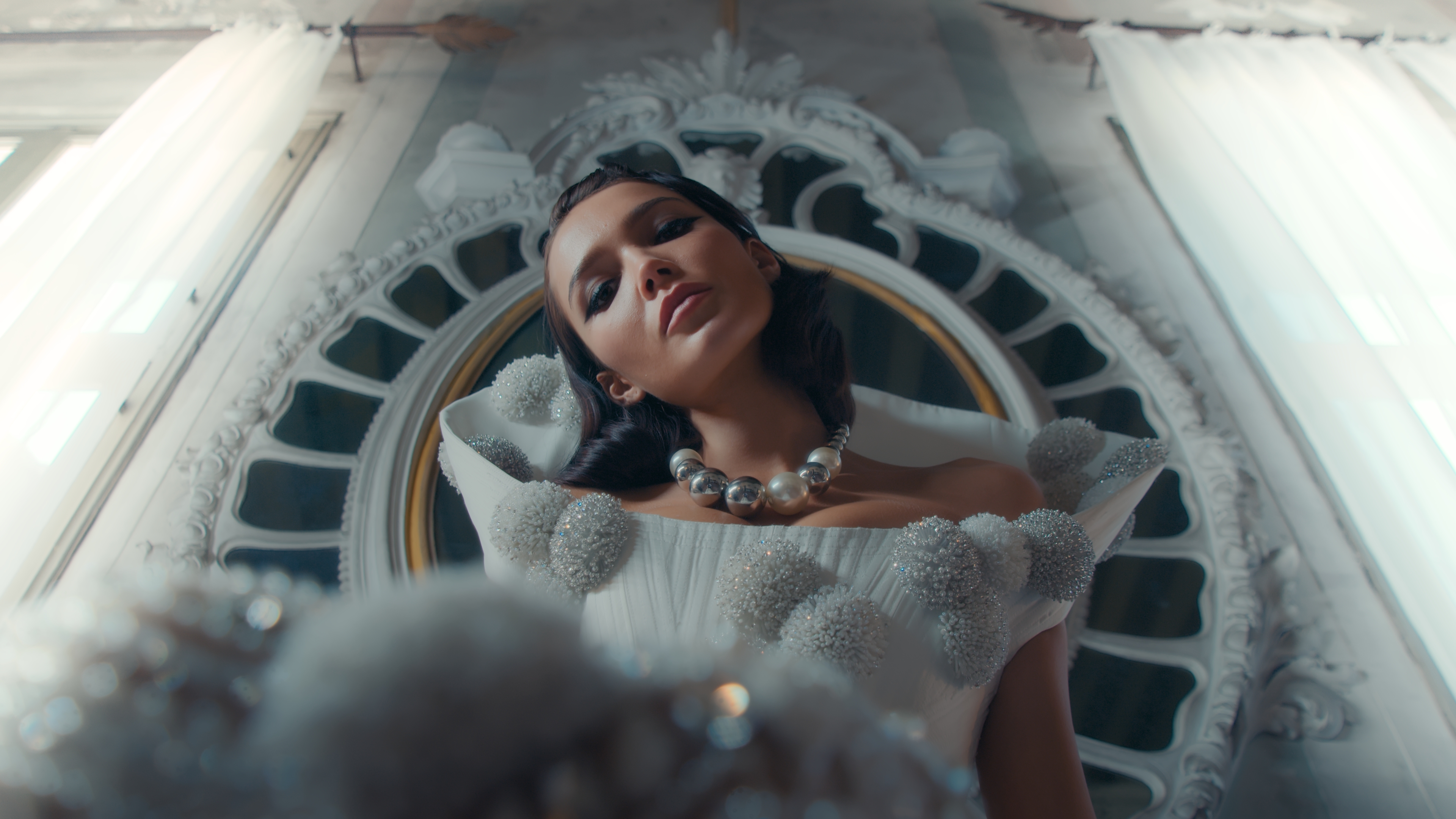 Elegant woman in white gown and pearl necklace poses in ornate indoor space with white walls and window light, fashion editorial.