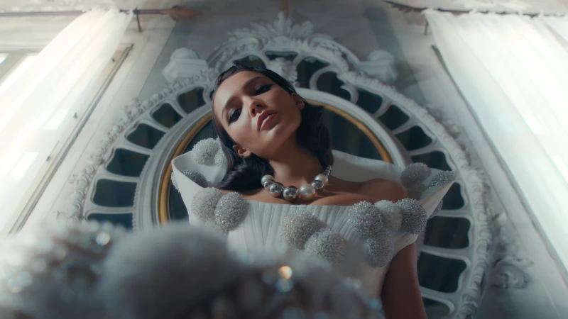 Elegant woman in white gown and pearl necklace poses in ornate indoor space with white walls and window light, fashion editorial.