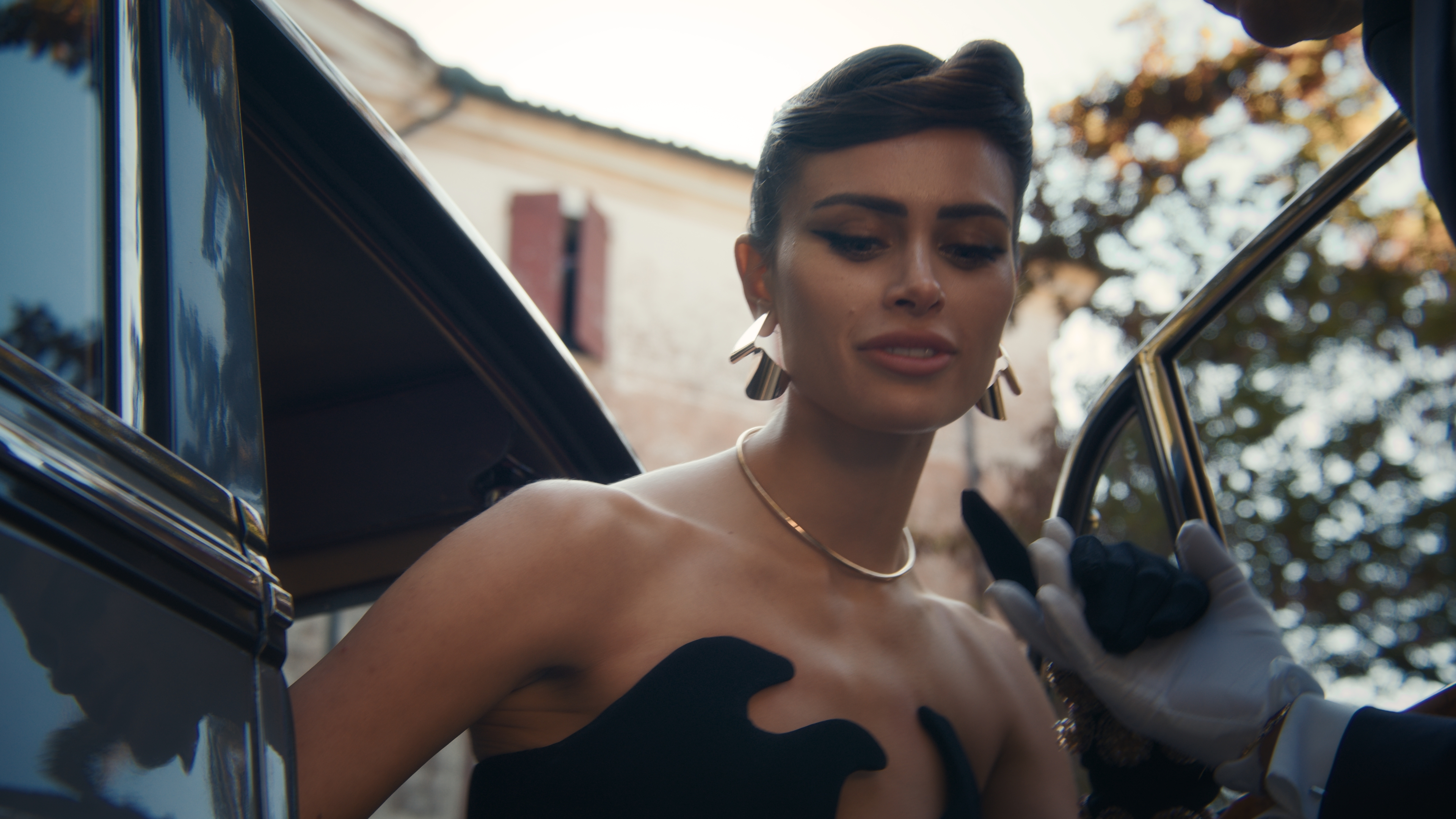 Elegant woman in a black dress steps out of a vintage car, assisted by a gloved attendant in an outdoor, sunlit setting.