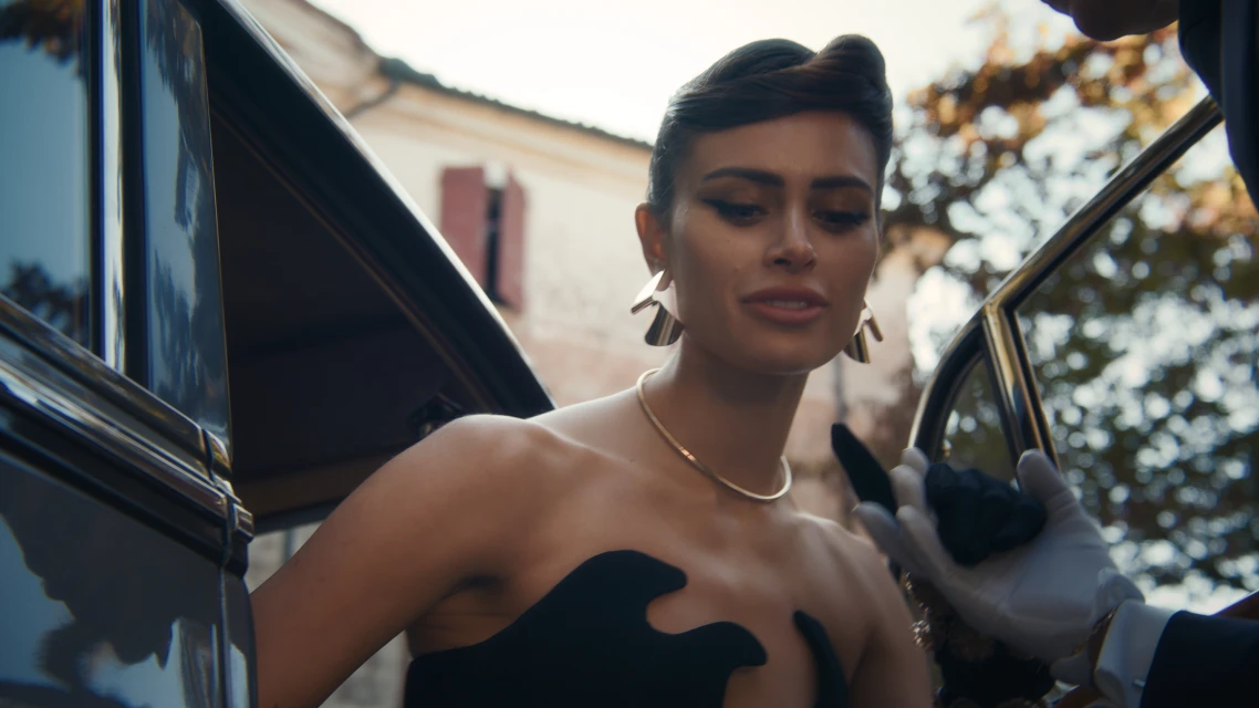 Elegant woman in a black dress steps out of a vintage car, assisted by a gloved attendant in an outdoor, sunlit setting.