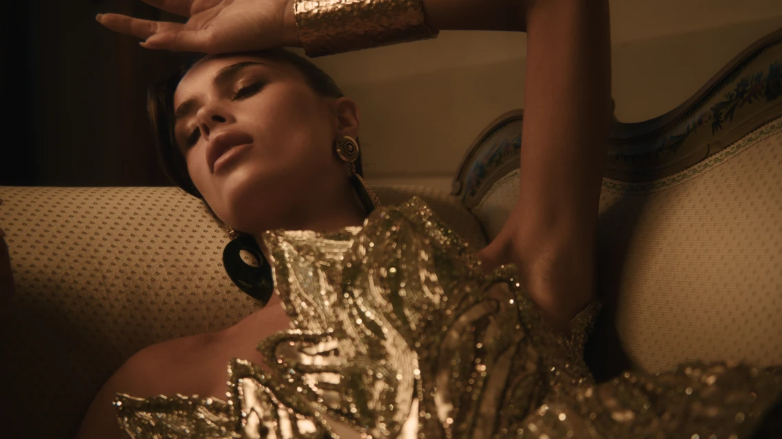 Attractive young woman lounges on an ornate couch wearing a gold dress and cuff. The indoor shot features warm tones and glamorous fashion.