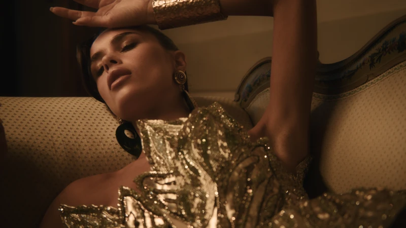 Attractive young woman lounges on an ornate couch wearing a gold dress and cuff. The indoor shot features warm tones and glamorous fashion.