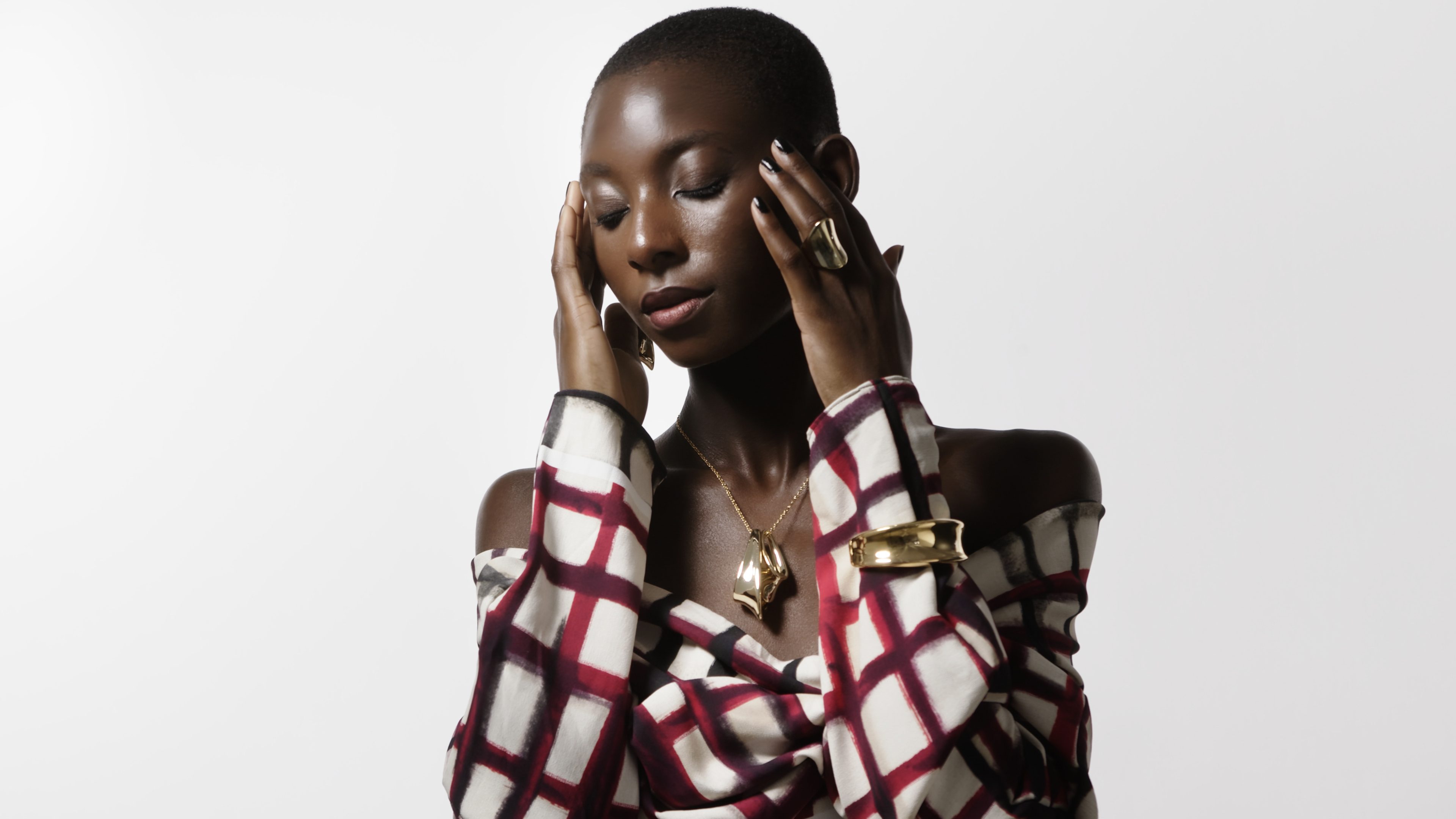Striking portrait of an African woman in a patterned top, gold jewelry, with hands near her face, eyes closed, against a bright white background.