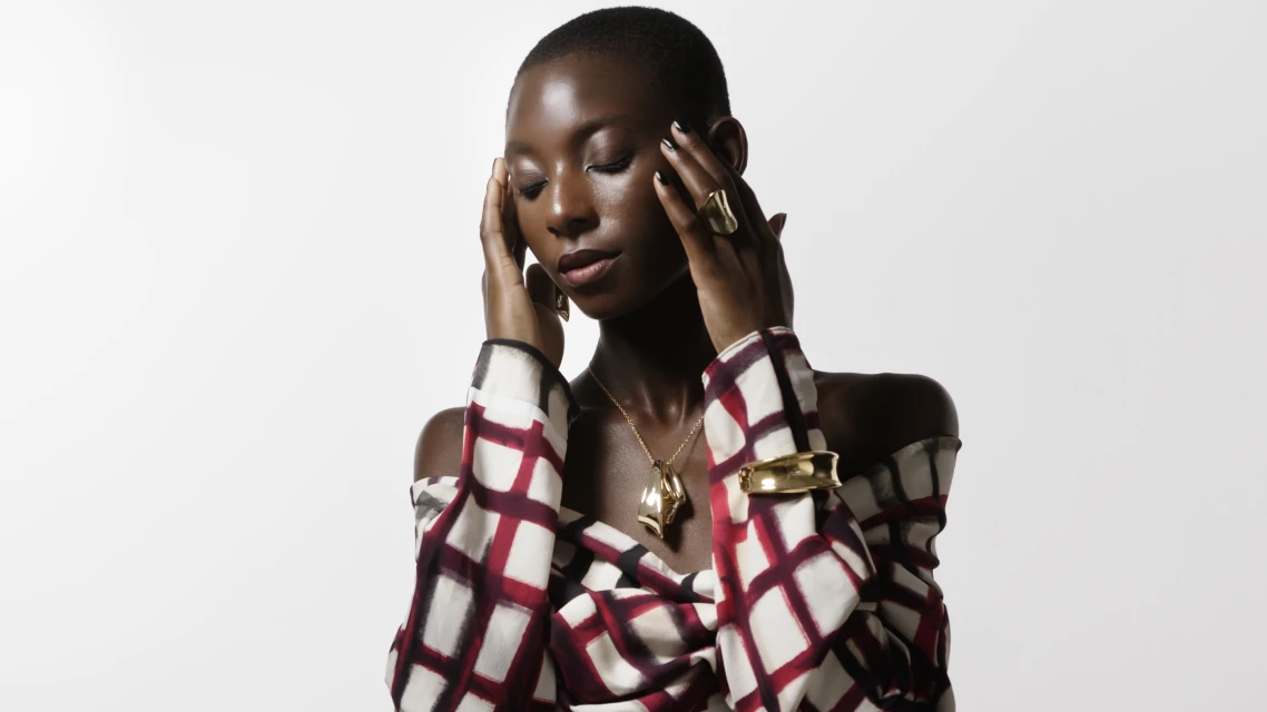 Striking portrait of an African woman in a patterned top, gold jewelry, with hands near her face, eyes closed, against a bright white background.
