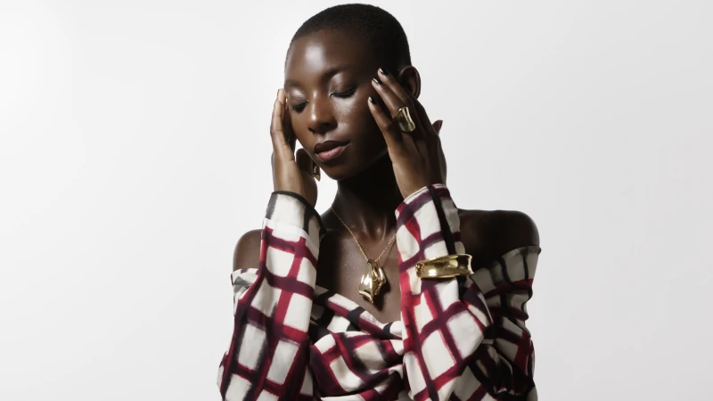 Striking portrait of an African woman in a patterned top, gold jewelry, with hands near her face, eyes closed, against a bright white background.