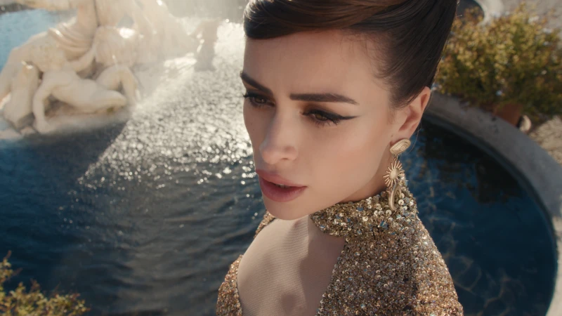 Woman with winged eyeliner and gold statement earring wearing a sequined gold dress in front of a stone fountain.