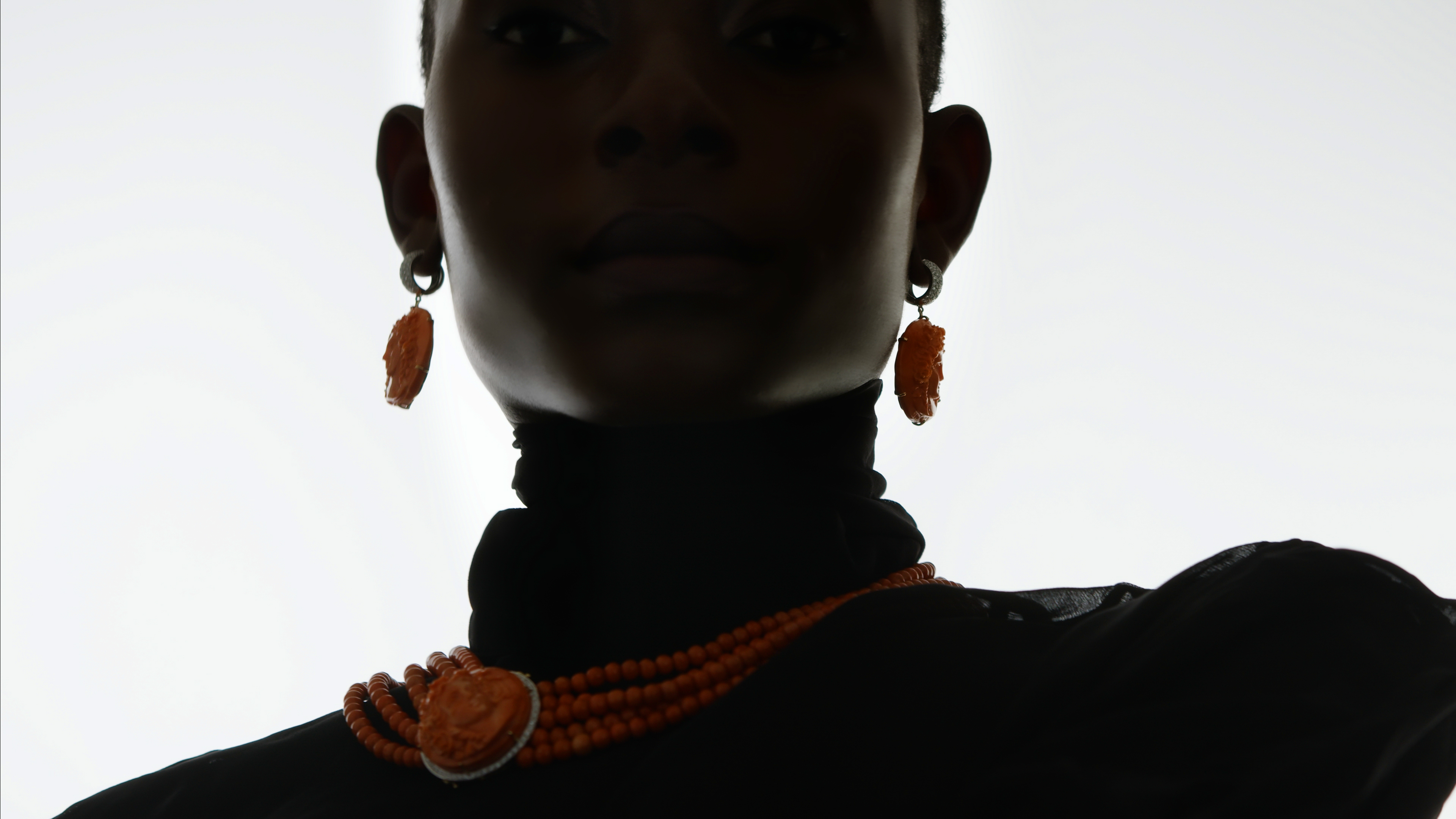 Close-up of a Black woman wearing orange beaded jewelry, including a necklace and earrings, against a bright, neutral background.