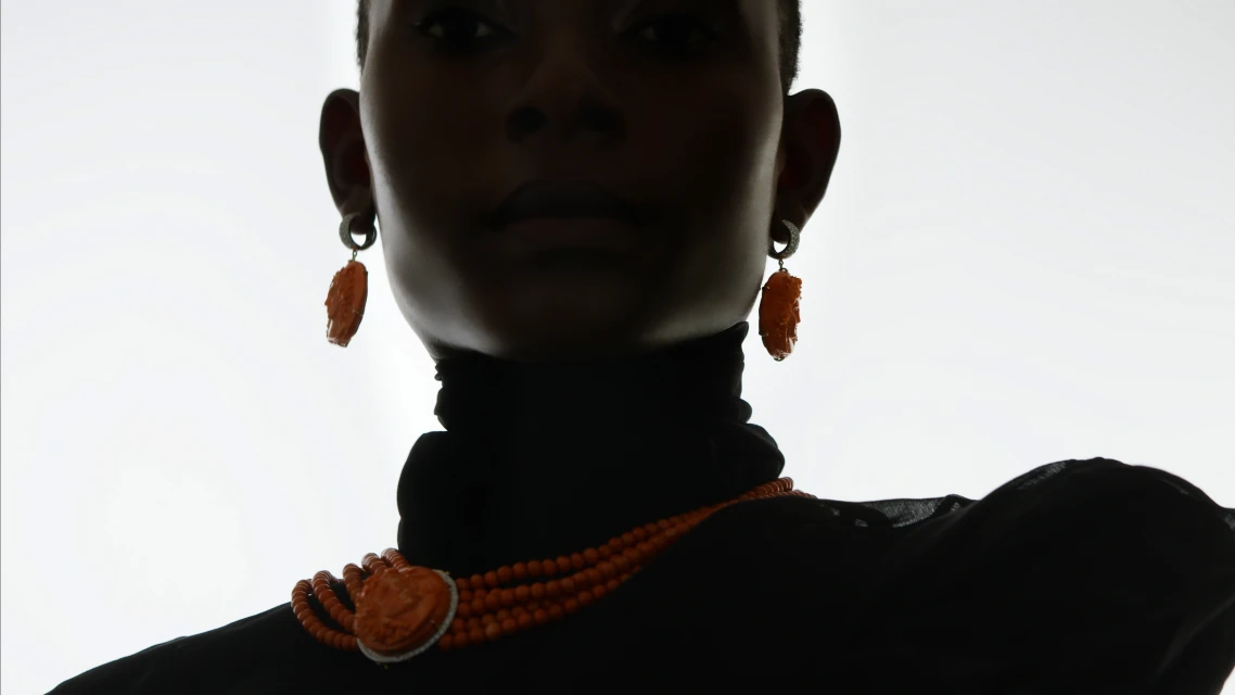 Close-up of a Black woman wearing orange beaded jewelry, including a necklace and earrings, against a bright, neutral background.