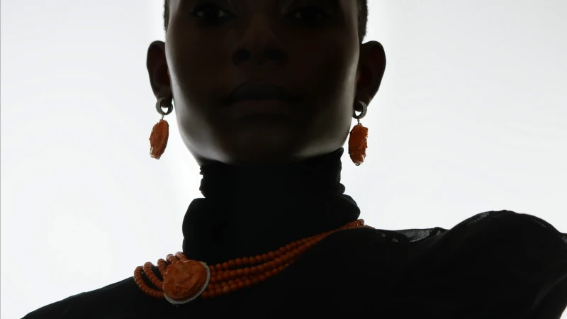 Close-up of a Black woman wearing orange beaded jewelry, including a necklace and earrings, against a bright, neutral background.