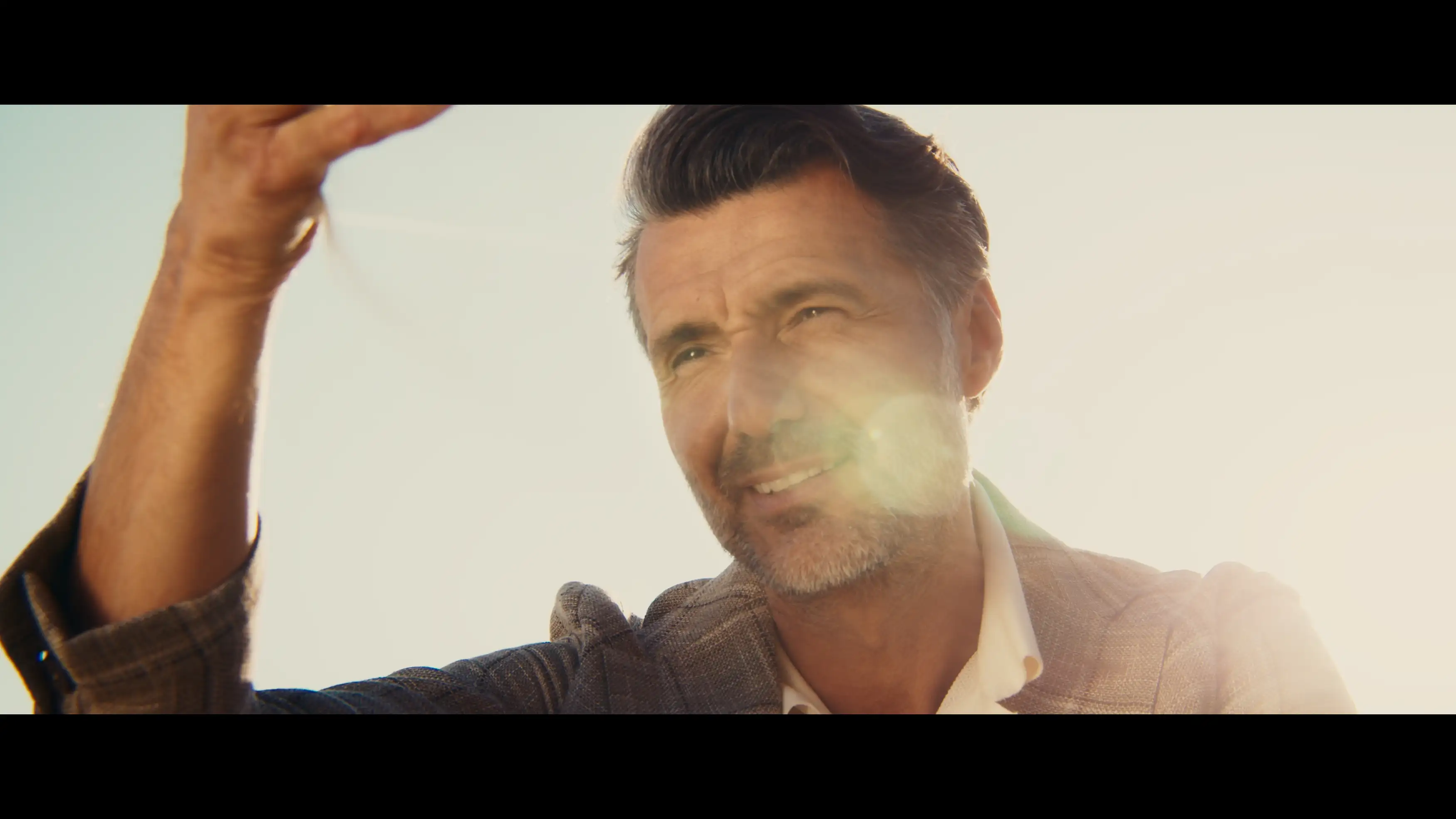 Close-up of a middle-aged Caucasian man in a patterned jacket, smiling slightly against a bright sky with a lens flare effect.