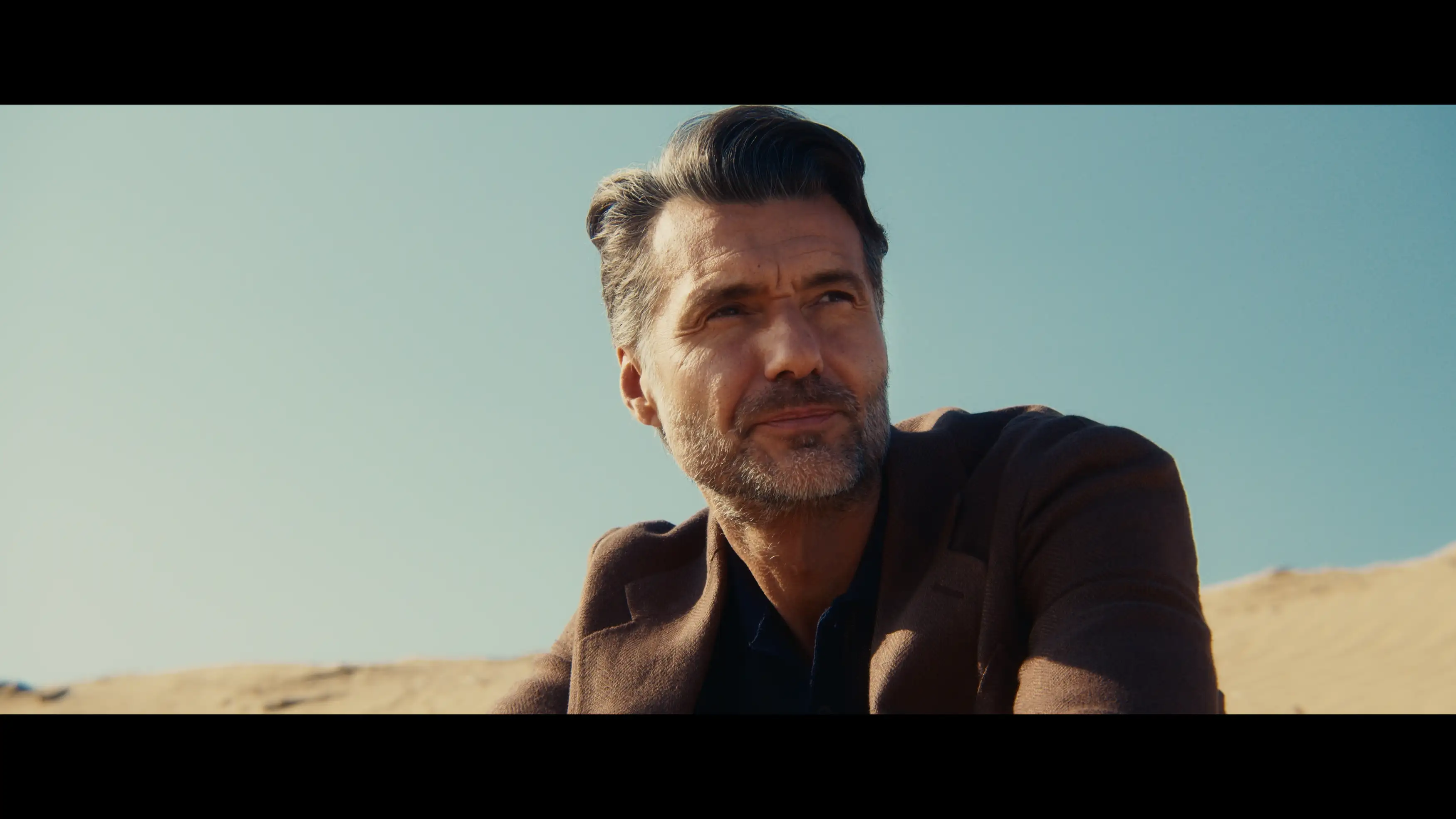 A middle-aged man in a tailored brown blazer looking thoughtfully into the distance against sunlit sand dunes.