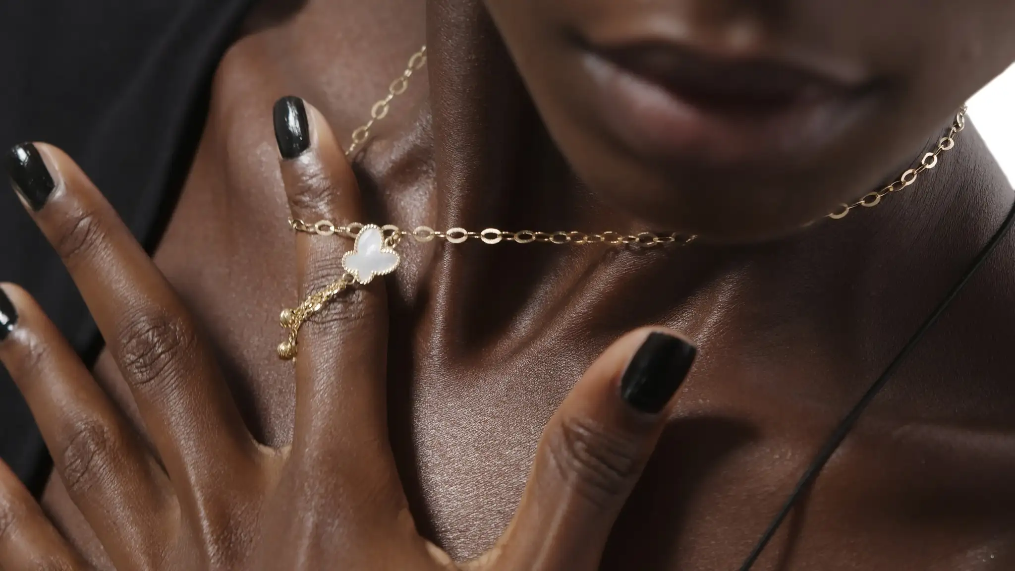 Close-up of a Black woman wearing a delicate gold chain necklace with a white pendant and black nail polish.