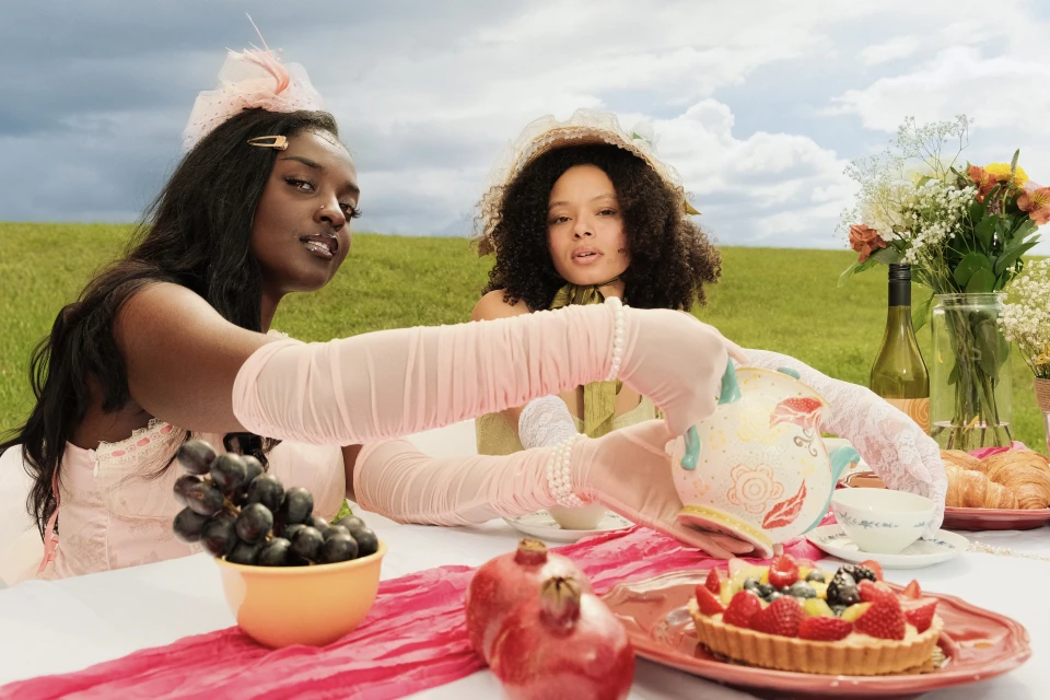 Two women enjoy an outdoor tea party. A table with tea, fruit, and flowers sits in a field under a cloudy sky, creating a charming scene.